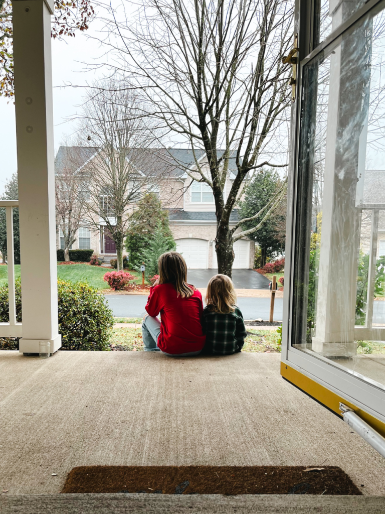 A girl and her younger sister sit together on the porch, looking toward the street.