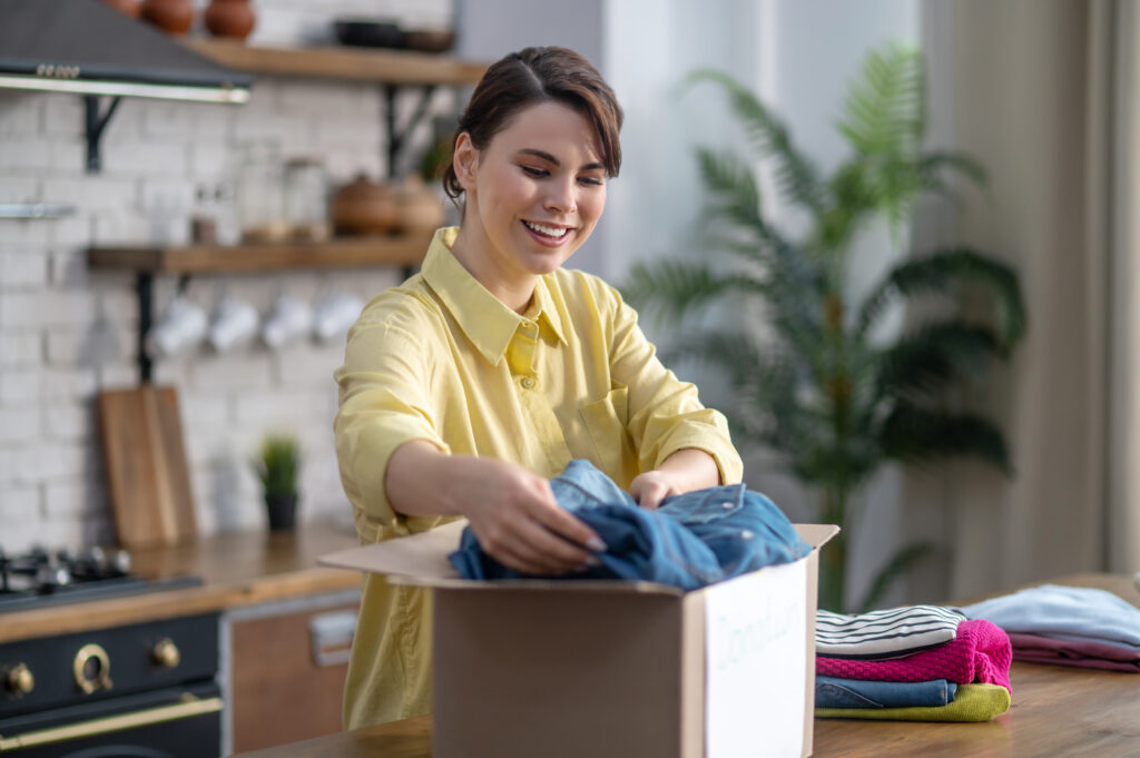 Joyful dark-haired woman packing folded items of clothing in the cardboard box